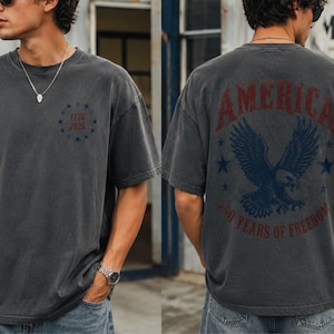 May include: A charcoal grey short-sleeve t-shirt featuring a small circular graphic on the front with the text 1776 2026 surrounded by stars. The back displays a large eagle graphic with the text AMERICA and 250 YEARS OF FREEDOM.