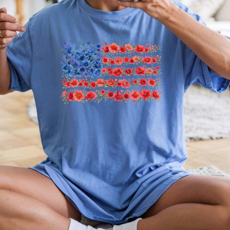 May include: A light blue t-shirt featuring a floral American flag design. The flag's stars are represented by blue flowers, and the stripes are made of red flowers. The shirt is a comfortable, casual style.