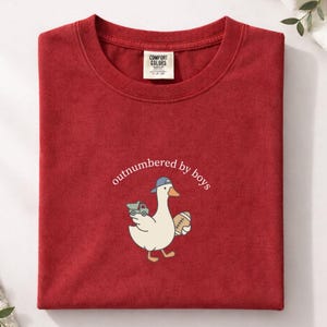 Op de afbeelding: Rood Comfort Colors T-shirt met een cartoon eend met een baseballpet en een voetbal en een speelgoed vrachtwagen. De tekst "outnumbered by boys" is boven de eend gebogen.
