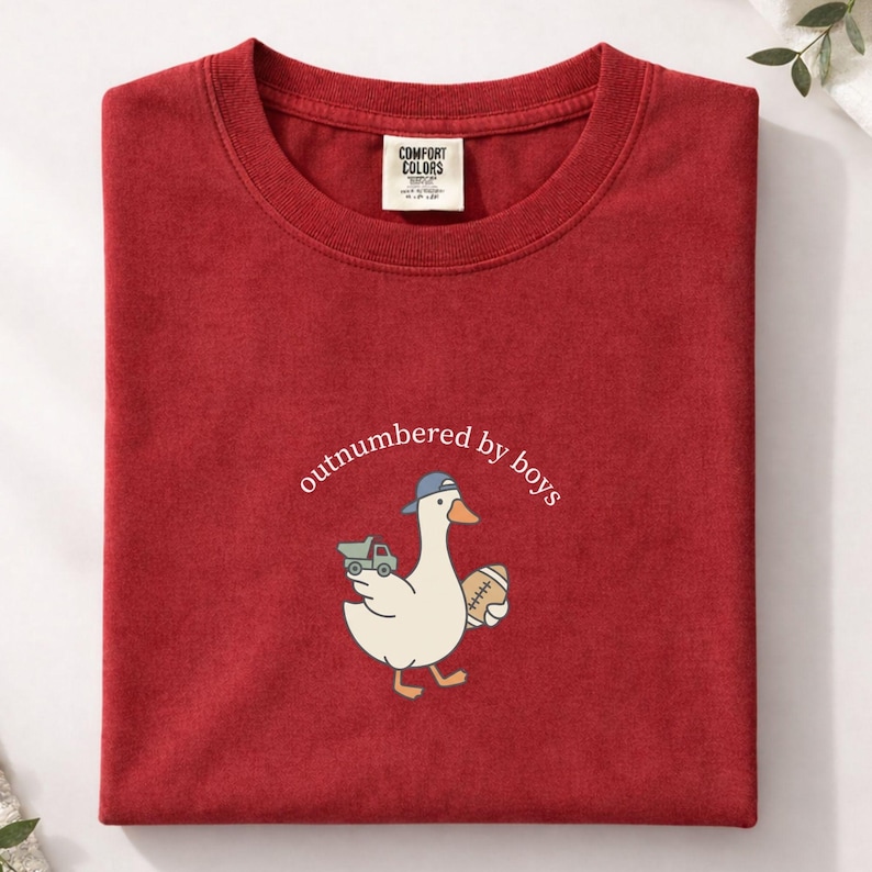 Op de afbeelding: Rood Comfort Colors T-shirt met een cartoon eend met een baseballpet en een voetbal en een speelgoed vrachtwagen. De tekst "outnumbered by boys" is boven de eend gebogen.