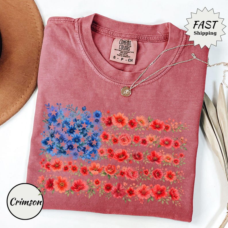 May include: A crimson-colored t-shirt featuring a floral American flag design. The flag's stars are represented by blue flowers, and the stripes are made of red and orange flowers. A silver necklace with a pendant is draped on the shirt.