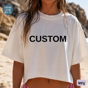 Op de afbeelding: Een ivoorkleurig, boxy t-shirt met korte mouwen en een ronde hals. Het woord CUSTOM is in dikke, zwarte hoofdletters in het midden op de borst gedrukt. Een cirkelvormige afbeelding in de linkerbovenhoek vermeldt COMFORT COLORS BOXY TEE.