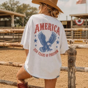 May include: A light gray t-shirt with a patriotic design. The back of the shirt features the word "AMERICA" in red, a blue eagle graphic, blue stars, and the text "250 YEARS OF FREEDOM".