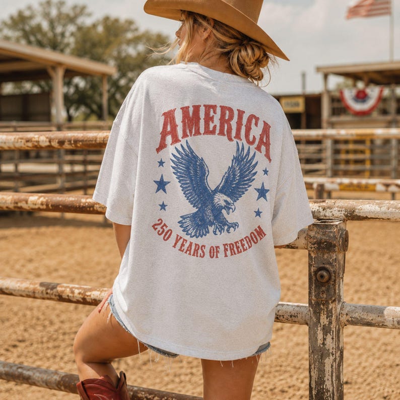 May include: A light gray t-shirt with a patriotic design. The back of the shirt features the word "AMERICA" in red, a blue eagle graphic, blue stars, and the text "250 YEARS OF FREEDOM".