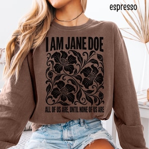 Op de afbeelding: Een espressobruine sweater met ronde hals en zwarte opdruk met de tekst I AM JANE DOE bovenaan en ALL OF US ARE, UNTIL NONE OF US ARE onderaan, voorzien van een centrale afbeelding van zwarte bloemranken en grote bloemen.