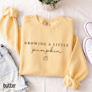 May include: A butter yellow crewneck sweatshirt featuring the text "GROWING A LITTLE pumpkin" in dark brown lettering, accompanied by a small line-art pumpkin icon centered below the text. The garment has long sleeves and a ribbed collar.