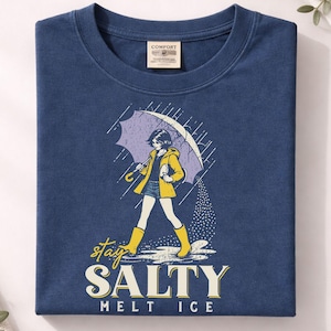 May include: Navy blue t-shirt featuring a graphic of a person walking in the rain under an umbrella. The person is wearing a yellow jacket and boots. The text "stay SALTY melt ICE" is below the graphic.