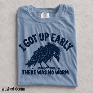 Könnte beinhalten: Ein kurzärmeliges T-Shirt in verwaschenem Denim-Blau mit einer dunkelblauen Grafik eines Raben. Der Text über dem Raben lautet I GOT UP EARLY und der Text darunter lautet THERE WAS NO WORM. Das T-Shirt hat einen Rundhalsausschnitt und ein Comfort Colors Etikett.
