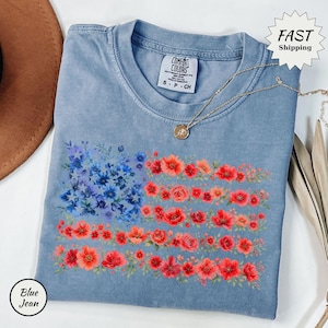 May include: A blue jean colored t-shirt featuring a floral American flag design. The flag's stars are represented by blue flowers, and the stripes are made of red and orange flowers. A gold necklace and a brown hat are also visible.