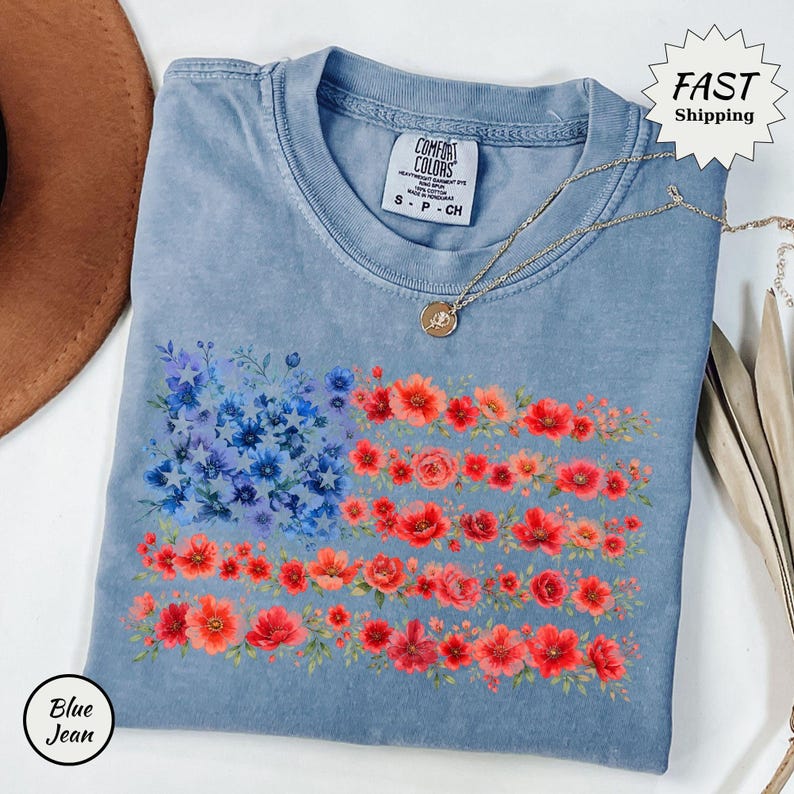 May include: A blue jean colored t-shirt featuring a floral American flag design. The flag's stars are represented by blue flowers, and the stripes are made of red and orange flowers. A gold necklace and a brown hat are also visible.