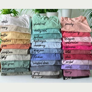 May include: A collection of folded t-shirts stacked in three columns, featuring a wide variety of colors labeled with text including white, ivory, butter, mustard, yam, sandstone, bay, light green, blue spruce, moss, island reef, chalky mint, seafoam, hydrangea, chambray, blue jean, flo blue, washed denim, gray, pepper, peachy, crunch berry, blossom, watermelon, heliconia, brick, chili, orchid, violet, berry, and black.