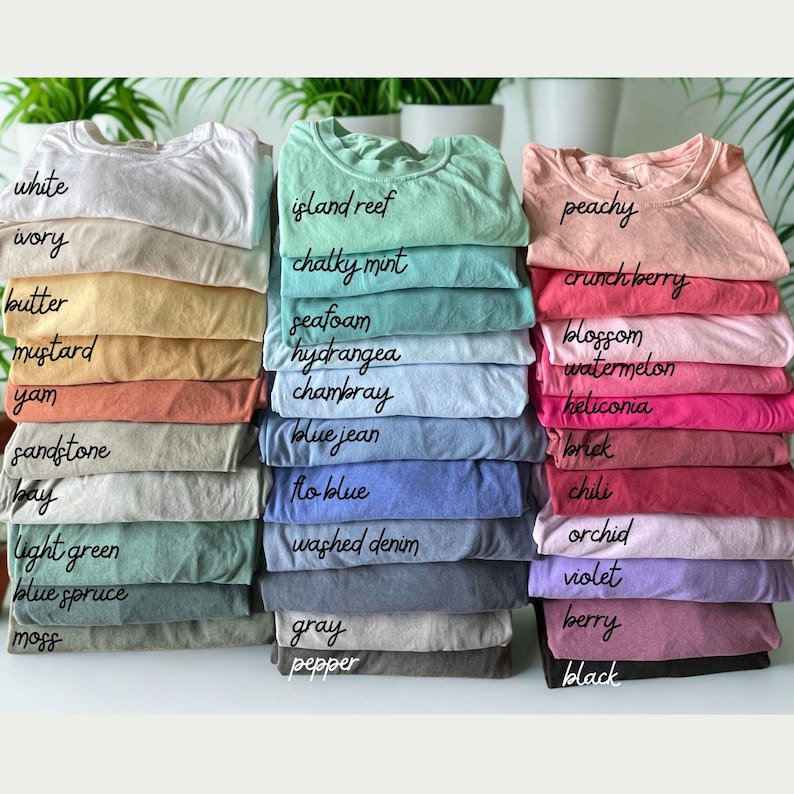 May include: A collection of folded t-shirts stacked in three columns, featuring a wide variety of colors labeled with text including white, ivory, butter, mustard, yam, sandstone, bay, light green, blue spruce, moss, island reef, chalky mint, seafoam, hydrangea, chambray, blue jean, flo blue, washed denim, gray, pepper, peachy, crunch berry, blossom, watermelon, heliconia, brick, chili, orchid, violet, berry, and black.