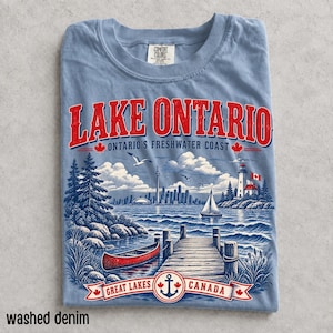 K&ouml;nnte beinhalten: Ein hellblaues T-Shirt mit einem Grafikdruck des Lake Ontario, der ein rotes Kanu, einen Holzsteg, einen Leuchtturm und eine Stadtsilhouette zeigt. Der Text lautet Lake Ontario, Ontario's Freshwater Coast und Great Lakes Canada mit roten Ahornblatt-Symbolen.