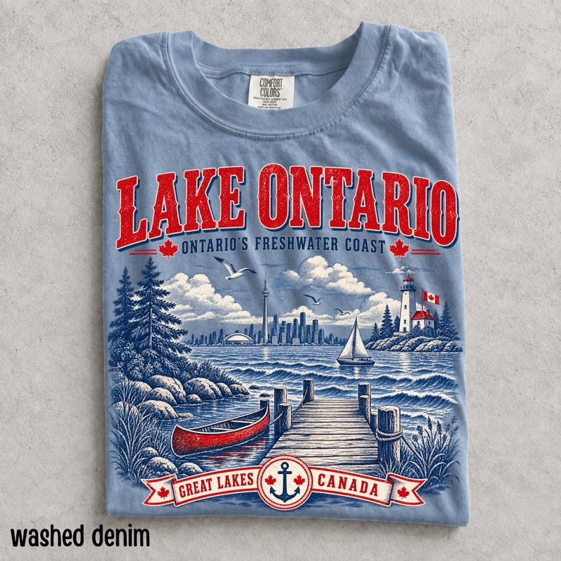 K&ouml;nnte beinhalten: Ein hellblaues T-Shirt mit einem Grafikdruck des Lake Ontario, der ein rotes Kanu, einen Holzsteg, einen Leuchtturm und eine Stadtsilhouette zeigt. Der Text lautet Lake Ontario, Ontario's Freshwater Coast und Great Lakes Canada mit roten Ahornblatt-Symbolen.