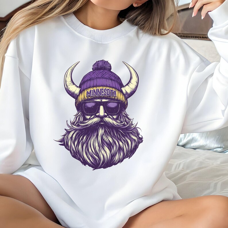 Pu&ograve; includere: Felpa bianca con grafica di un vichingo con berretto in maglia viola e giallo con la scritta "MINNESOTA", occhiali da sole e lunga barba viola. Il vichingo ha grandi corna ricurve. La felpa &egrave; a girocollo e maniche lunghe.