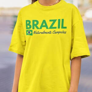 May include: A yellow t-shirt with the word "BRAZIL" in green block letters. Below the word is the Brazilian flag and the words "Naturalmente Campe&otilde;es" in green script.