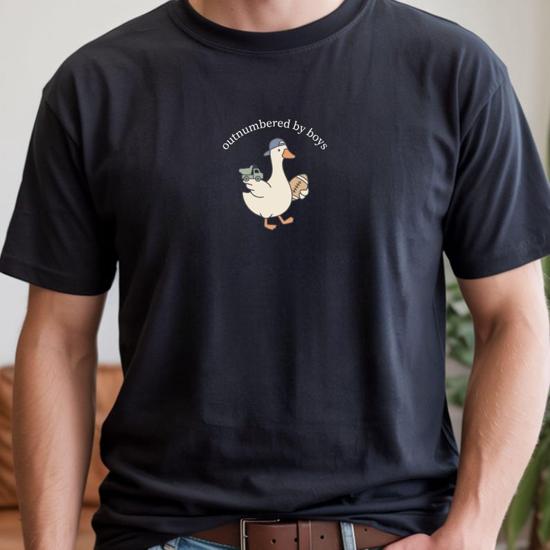 Op de afbeelding: Zwart t-shirt met een cartoon gans met een pet en een speelgoed vrachtwagen en een rugbybal. De tekst "outnumbered by boys" staat boven de gans. Het shirt is gemaakt van een zacht materiaal.