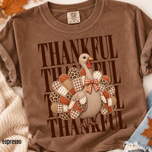 Puede incluir: Una camiseta marrón de manga corta con el gráfico de un pavo con una cola de patchwork hecha de estampados de leopardo, cuadros y lunares. El pavo lleva un lazo de cuadros rojos y blancos. La palabra THANKFUL se repite en el fondo del diseño.