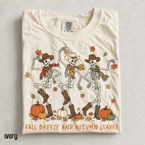 May include: An ivory t-shirt featuring a graphic of three dancing skeletons wearing cowboy hats and scarves. The skeletons are surrounded by falling autumn leaves and pumpkins. The text at the bottom reads, Fall Breeze and Autumn Leaves.