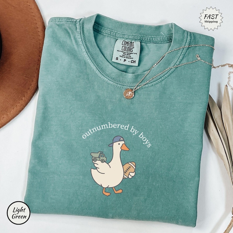 Op de afbeelding: Een lichtgroen t-shirt met de tekst "outnumbered by boys" en een cartoon eend met een baseballpet en een voetbal. Het shirt heeft ook een kleine vrachtwagenafbeelding. Een zilveren ketting met een hanger is over het shirt gedrapeerd.