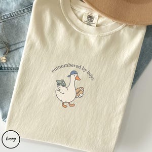 Op de afbeelding: Ivoor kleurig T-shirt met de tekst "outnumbered by boys" en een cartoon gans met een baseballpet en een speelgoed vrachtwagen en voetbal. Het shirt heeft een ronde hals en een Comfort Colors label.