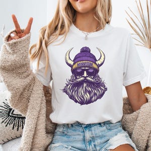 Pu&ograve; includere: T-shirt bianca con grafica di un vichingo barbuto con un berretto di lana viola e oro con la scritta "MINNESOTA". Il vichingo ha occhiali da sole e corna. Il design &egrave; nelle tonalit&agrave; del viola e dell'oro.