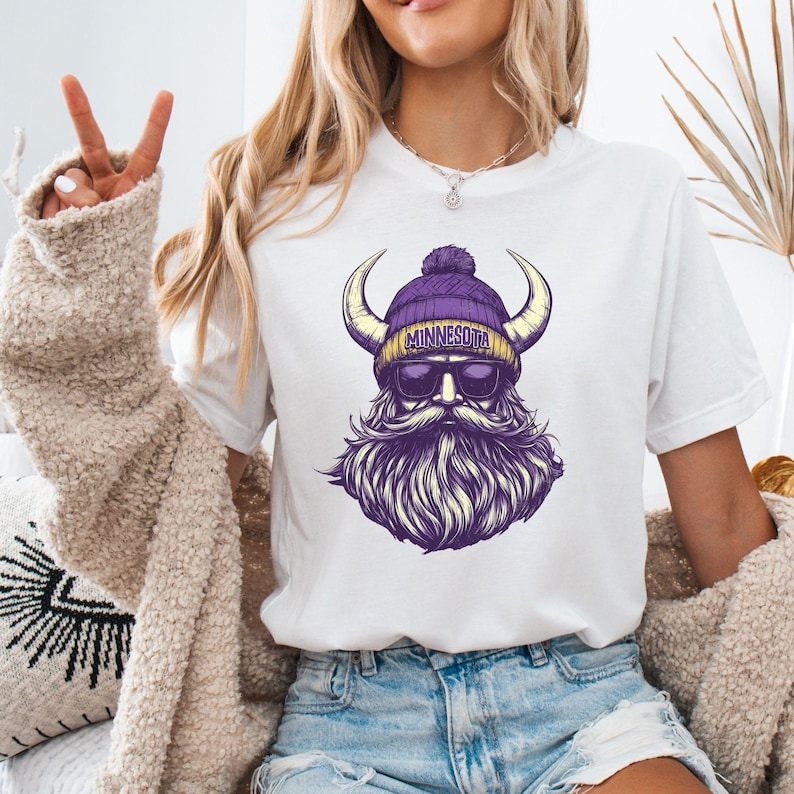 Pu&ograve; includere: T-shirt bianca con grafica di un vichingo barbuto con un berretto di lana viola e oro con la scritta "MINNESOTA". Il vichingo ha occhiali da sole e corna. Il design &egrave; nelle tonalit&agrave; del viola e dell'oro.