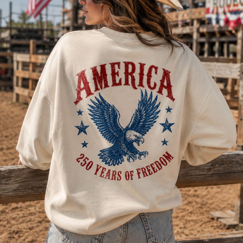May include: Cream-colored sweatshirt with a distressed red "AMERICA" graphic above a blue eagle illustration. Below the eagle, the text reads "250 YEARS OF FREEDOM" in red. The design is patriotic and vintage-inspired.