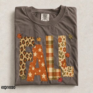 Puede incluir: Una camiseta de manga corta gris oscuro con la palabra FALL en letras grandes. Cada letra tiene un patrón único: estampado de leopardo, naranja con estrellas blancas, cuadros verdes y naranjas, y un estampado floral con flores naranjas.