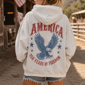 May include: A light gray hoodie with the word "AMERICA" in red, arched across the top. Below is a blue eagle with outstretched wings, surrounded by blue stars. The text "250 YEARS OF FREEDOM" is below the eagle.