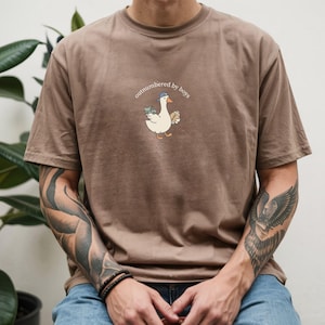 Op de afbeelding: Bruin t-shirt met de tekst "outnumbered by boys" en een cartoon gans met een hoed, geld en een sandwich. Het shirt is gemaakt van een zacht materiaal en heeft korte mouwen.
