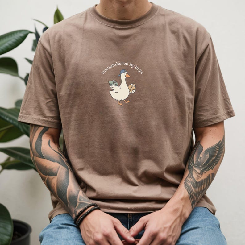 Op de afbeelding: Bruin t-shirt met de tekst "outnumbered by boys" en een cartoon gans met een hoed, geld en een sandwich. Het shirt is gemaakt van een zacht materiaal en heeft korte mouwen.