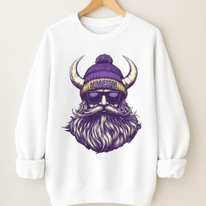 Pu&ograve; includere: Felpa bianca con grafica di un vichingo barbuto con berretto viola con la scritta "MINNESOTA" e occhiali da sole. Il vichingo ha grandi corna e una lunga barba stilizzata. La felpa ha maniche lunghe.