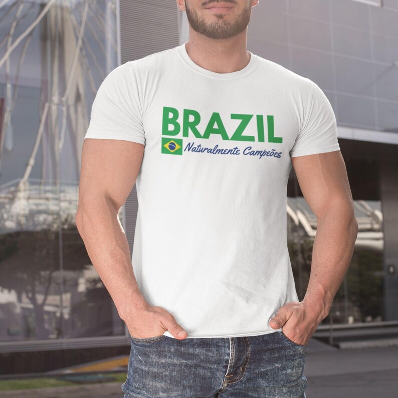 May include: White t-shirt with the word "BRAZIL" in green block letters, a small Brazilian flag, and the text "Naturalmente Campe&otilde;es" in blue. The shirt is worn by a person in blue jeans.