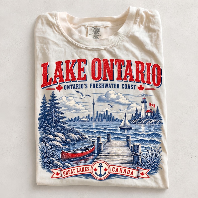 K&ouml;nnte beinhalten: Ein cremefarbenes T-Shirt mit einer Grafik des Lake Ontario. Das Design zeigt ein rotes Kanu, einen Holzsteg, einen Leuchtturm, ein Segelboot und die Skyline von Toronto. Der Text lautet LAKE ONTARIO, ONTARIO'S FRESHWATER COAST und GREAT LAKES CANADA.