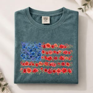 May include: A teal Comfort Colors t-shirt folded neatly. The front features a floral American flag design, with blue flowers forming the stars and red and orange flowers creating the stripes. The shirt has a crew neck and a small tag.