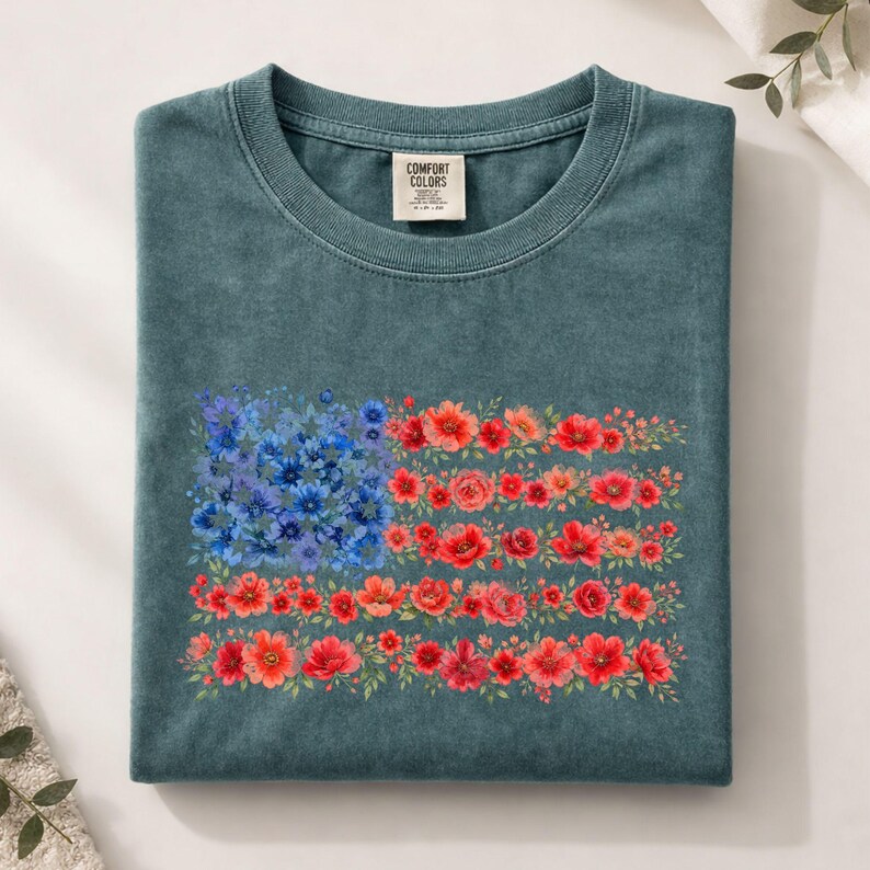 May include: A teal Comfort Colors t-shirt folded neatly. The front features a floral American flag design, with blue flowers forming the stars and red and orange flowers creating the stripes. The shirt has a crew neck and a small tag.