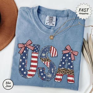 Op de afbeelding: Een blauw spijkerbroek-T-shirt met het woord "USA" in een patriottisch ontwerp. De letters hebben een combinatie van Amerikaanse vlagpatronen, luipaardprint en rode strikken. Een gouden ketting en een bruine hoed zijn ook zichtbaar.