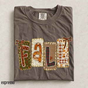 May include: A dark brown crew neck t-shirt featuring the word FALL in large block letters. Each letter has a unique pattern including pumpkins, plaid, and woodland animals, with decorative bows on the F and L. The shirt has a Comfort Colors label.