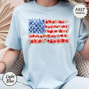 May include: Light blue t-shirt featuring a floral American flag design. The flag's stars are represented by blue flowers, and the stripes are red flowers. The shirt also has the text "Light Blue" and "Fast Shipping".