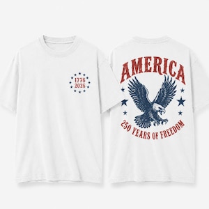 May include: A white t-shirt featuring a front graphic of the text 1776 2026 surrounded by a circle of blue stars. The back graphic displays a large eagle with the text AMERICA 250 YEARS OF FREEDOM in red and blue with a distressed texture.