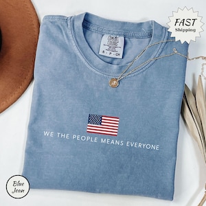 Puede incluir: Una camiseta de color azul vaquero con cuello redondo y una etiqueta de la marca Comfort Colors en el cuello. El centro de la camiseta presenta un pequeño gráfico de la bandera estadounidense sobre el texto blanco, WE THE PEOPLE MEANS EVERYONE.