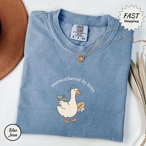 Op de afbeelding: Blauw spijkerbroekkleurig T-shirt met de tekst "outnumbered by boys" en een cartoon eend met een baseballpet en een voetbal en een speelgoedvrachtwagen. Een gouden ketting is over het shirt gedrapeerd.