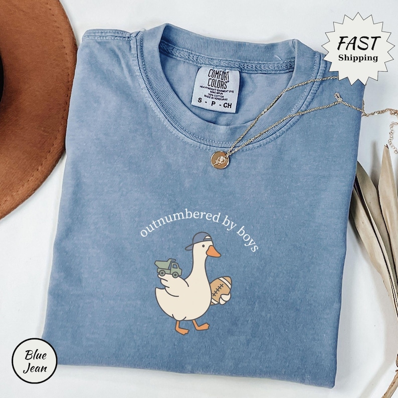 Op de afbeelding: Blauw spijkerbroekkleurig T-shirt met de tekst "outnumbered by boys" en een cartoon eend met een baseballpet en een voetbal en een speelgoedvrachtwagen. Een gouden ketting is over het shirt gedrapeerd.