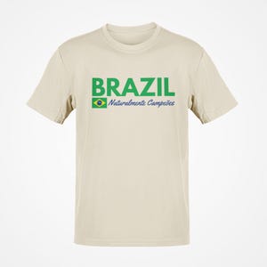 May include: A beige t-shirt with the word "BRAZIL" in green block letters. Below the word "BRAZIL" is a small Brazilian flag and the words "Naturalmente Campe&otilde;es" in blue. The t-shirt is a crew neck style.