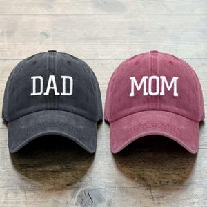 Puede incluir: Dos gorras de béisbol, una gris oscura y otra burdeos. La gorra gris oscura tiene la palabra "DAD" bordada en blanco, y la gorra burdeos tiene "MOM" bordado en blanco. Ambas gorras tienen una visera curva y una correa ajustable.