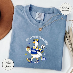 Può includere: T-shirt blu jeans Comfort Colors con grafica di un'anatra in divisa da calcio bosniaca, con caffè e la scritta "Off To Watch Soccer 2026". Sono presenti anche un pallone da calcio e delle stelle.