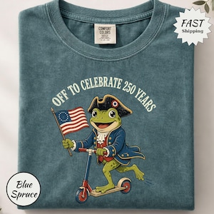Könnte beinhalten: Blaugrünes T-Shirt mit einem Frosch, der Roller fährt und eine Flagge hält. Der Frosch trägt einen Dreispitz und einen blauen Mantel. Der Text lautet "OFF TO CELEBRATE 250 YEARS". Das T-Shirt ist gefaltet.