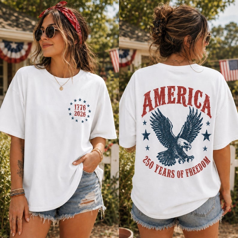 May include: A white short-sleeve t-shirt featuring a small circular graphic on the front chest with blue stars and the text 1776 2026. The back displays a large graphic of a bald eagle with the text AMERICA 250 YEARS OF FREEDOM in red and blue.