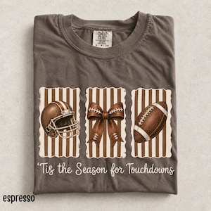 以下が含まれることがあります： ダークブラウンのTシャツで、茶色と白のストライプが入った3つの縦長長方形パネルが描かれています。パネルにはフットボール用ヘルメット、リボン、フットボールが収められています。グラフィックの下には白い文字で「Tis the Season for Touchdowns」とプリントされています。